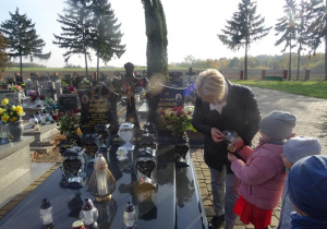 Dziewczynka trzyma w ręku znicz, który podpala pani Arletka. Stoją przy grobach księdza Bogusława Palenickiego oraz księdza Ryszarda Kolibskiego, w tle inne groby.